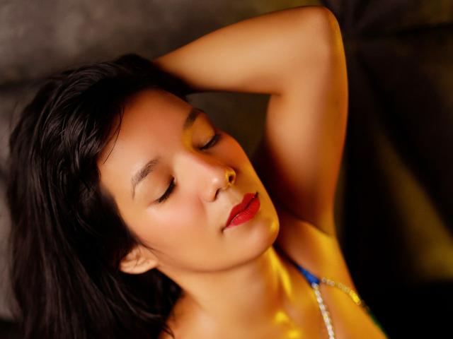 Close-up of LuciaMoretti with dark, wavy hair, eyes closed, and red lipstick. She has a small nose stud on her right nostril. Her right arm is raised, with her hand resting on the back of her head. She wears a blue and white necklace. Warm, yellow light illuminates her face, highlighting smooth skin and a relaxed expression. The background is dark and out of focus. The image has a soft, almost cinematic quality.