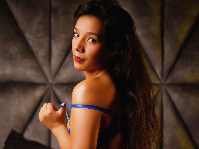 LuciaMoretti has long, dark brown hair flowing over her shoulders. She's looking over her shoulder with a slight smile, her brown eyes focused on the camera. She's holding a blue strap, which is part of a revealing outfit. Her skin is smooth and has a warm, light brown tone. The lighting highlights her facial features and creates a subtle shadow on the right side of her face. Her red lipstick adds a pop of color to her face. The background is a dark, textured wall with a geometric pattern. The composition is centered and close-up, focusing on LuciaMoretti's upper body and face.