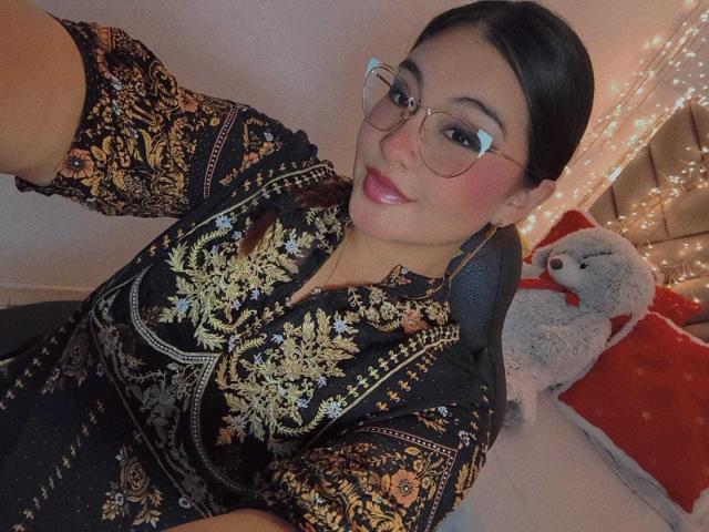 HelueJamil with glasses, black hair in a ponytail, wearing an ornate black dress with gold embroidery, sitting on a chair, smiling, holding a white stuffed animal.