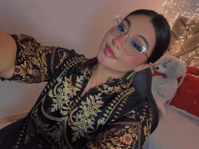 HelueJamil with glasses, dark hair in a ponytail, wearing a black floral shirt, smiling at the camera, sitting in a chair, with a stuffed gray dog in the background.