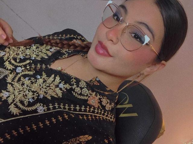 HelueJamil with braids, glasses, and pink lipstick, wearing a black embroidered top, sitting in a chair, looking at the camera with a slight pout, showing her tits.