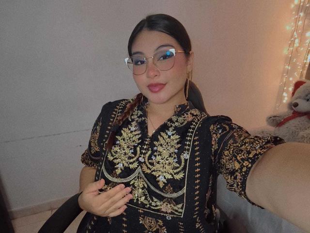 HelueJamil with glasses, dark hair, and red lipstick, wearing an ornate black dress with gold embroidery, holding her tits, sitting in a chair, and smiling.