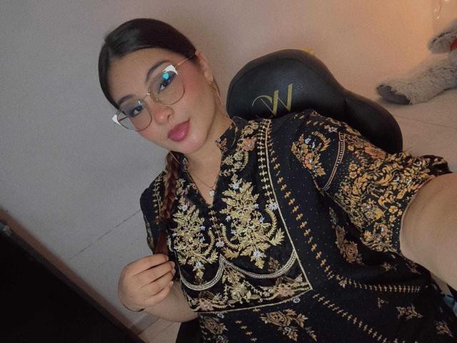 HelueJamil with glasses, dark hair in braid, wearing an ornate black and gold patterned top, sitting in a black chair, holding top's collar, smiling, pink lips, soft lighting.