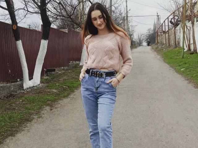 FortuneX with long brown hair, pink sweater, blue jeans, black belt, hands in pockets, standing on a gravel road, trees with white trunks, and a red fence.