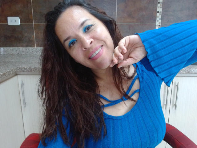 LizethBells, a medium-build woman with long, wavy brown hair, smiles at the camera. She wears a bright blue, ribbed, long-sleeve top with strappy details across the chest. Her blue eyeliner matches her top, creating a bold contrast. She rests her right hand on her cheek. The kitchen background has brown tiled walls and white cabinets with a red chair partially visible. The photo is clear with natural lighting.
