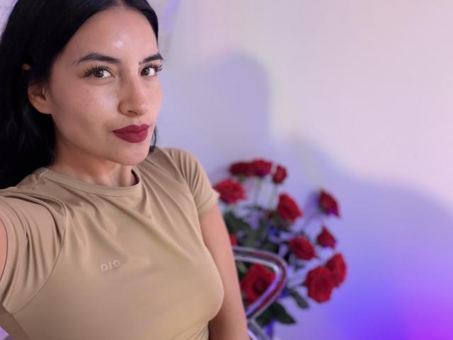 EmilyJensenn with long black hair, tan shirt, and dark red lipstick, smiling at camera, showing her tits, with a bouquet of red roses in the background.