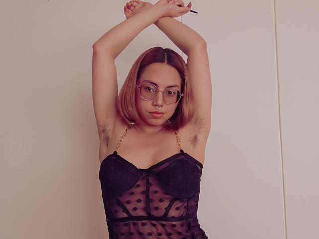 NaturalMariaHun with arms raised, showing armpit hair, wearing a black sheer polka dot top, glasses, and nose ring, looking at the camera. Her tits are visible through the top.