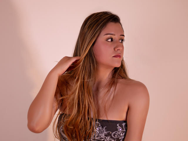 MiiaStark with long brown hair, touching her head, looking to the side, wearing a strapless grey top with purple patterns, soft pink skin, slight blush, subtle makeup.