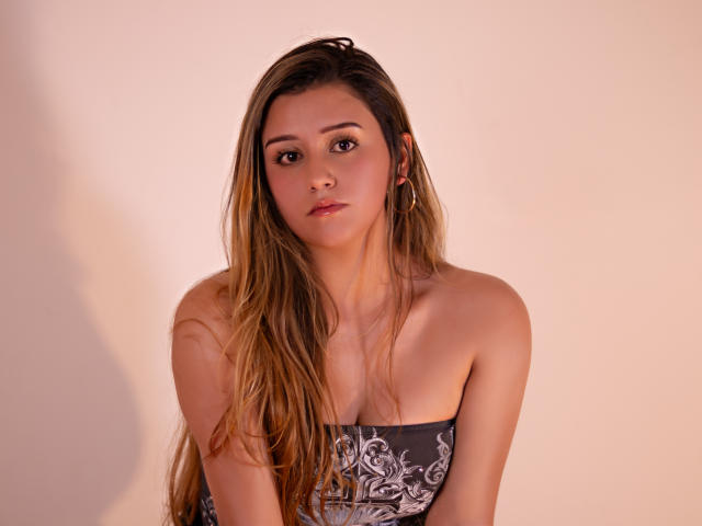 MiiaStark with long brown hair, wearing a strapless grey patterned top, has small tits and a neutral expression, standing against a soft pink wall.