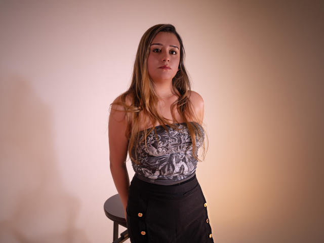 MiiaStark with long blonde hair, wearing a strapless grey top with black and gold buttoned skirt, standing against a beige wall, hands on a stool, looking slightly bored.