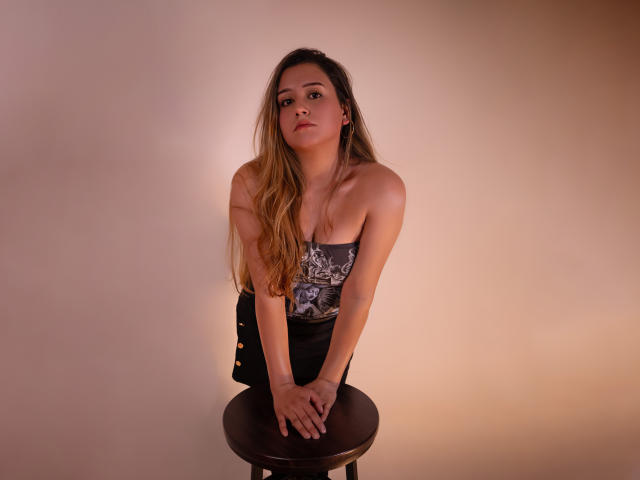 MiiaStark with long blonde hair, small tits, wearing a strapless gray top, leaning forward on a dark wooden stool, hands on seat, soft pink lighting.