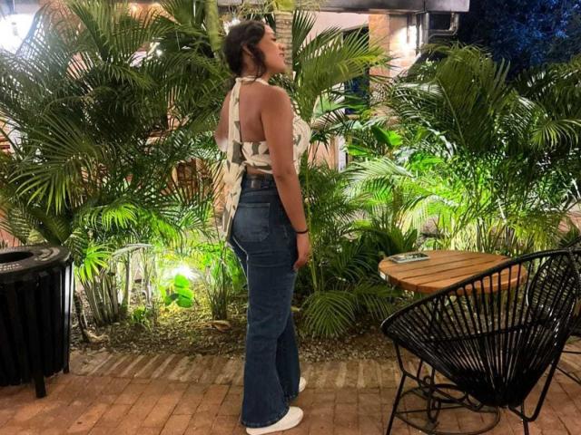 AraFerrer stands in a lush, green, tropical-style garden. She's facing away, showing her back. She wears a white halter-top with a black leaf pattern, and high-waisted, blue, flared jeans. Her dark hair is tied up. She's wearing white sneakers. The black, metal, round chair is on the right, and a black trash can is on the left. The ground is brick-paved. The image is lit with warm, greenish lighting.