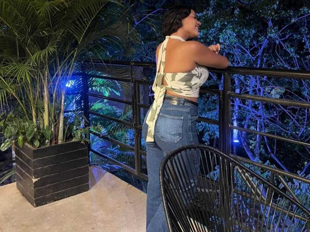 AraFerrer stands on a balcony at night, facing away. She wears a sleeveless, camouflage-patterned top and high-waisted blue jeans. Her arms rest on a black metal railing. She has short, dark hair. The balcony has a black, rectangular planter with green plants on the left. A curved black chair is in the lower right. Blue lights illuminate the dense, dark green foliage behind her. The floor is a light beige. The image has a casual, modern style.