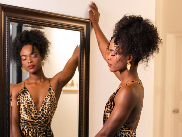 BelyiSinisterra stands in front of a mirror, her right hand touching the frame. She's wearing a leopard-print dress with a deep V-neck, gold earrings, and has curly black hair. Her reflection is clear in the mirror. She has dark skin, pink eyeshadow, and red lipstick. The mirror has a dark, wooden frame. Her expression is calm, and she's slightly turned to the right, showing her profile. The lighting is soft, highlighting her features and the texture of her dress.