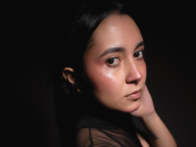 Gaby Mathew mit strahlender Haut, schwarzen Haaren, braunen Augen, einem Nasenring und goldenen Ohrringen, die ihre Wange berührt, blickt in die Kamera und trägt ein durchsichtiges schwarzes Oberteil vor einem dunklen Hintergrund..