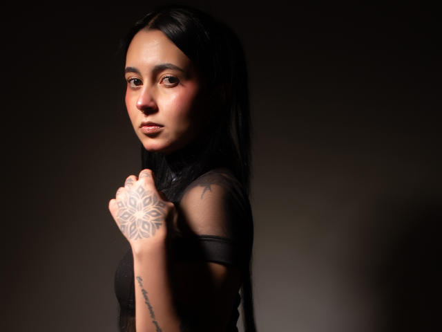 GabyMathew with long black hair, brown eyes, and a tattoo on her right hand, wearing a sheer black top, looking over her shoulder, holding her hand near her face.