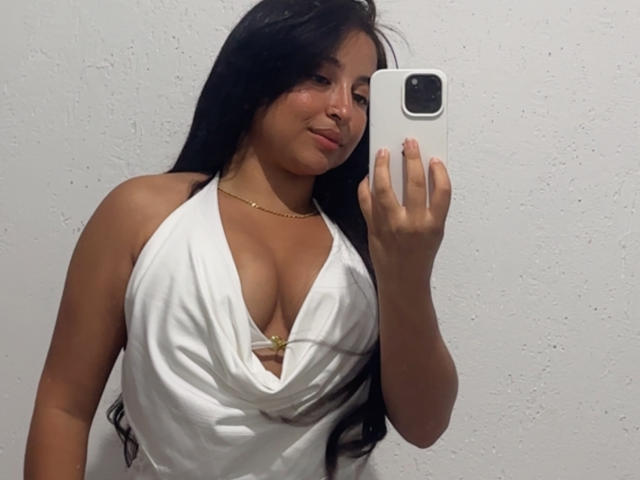 LarissaCruz with long black hair, wearing a deep white halter dress showing her big boobs, holding a white phone, gold necklace, and a subtle smile.