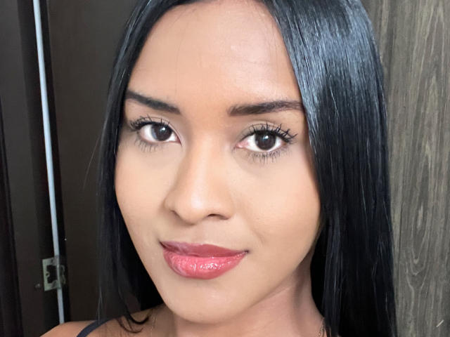 Close-up of Emyii, a woman with medium brown skin and straight black hair. She has dark brown eyes, thick black eyebrows, and long eyelashes. Her lips are full and glossy with pink lipstick. She's wearing a black strap, visible on the left. The image is sharply focused on her face, with a neutral brown background and a wooden texture on the right. Her expression is calm and slightly smiling. The lighting is even, highlighting her facial features.