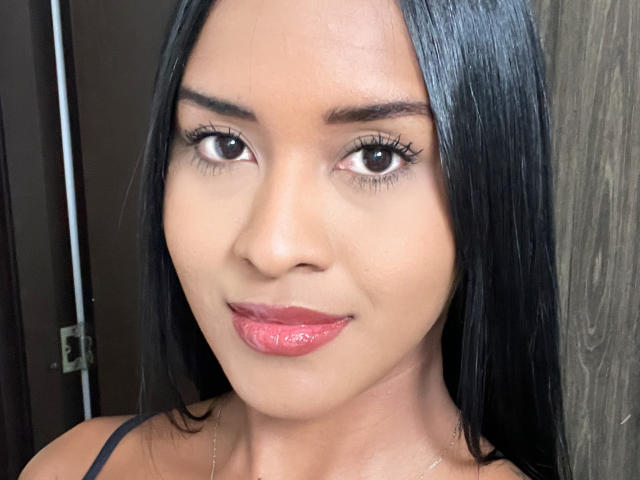 Close-up of Emyii with straight black hair, brown eyes, and pink lipstick. She has a smooth, tan complexion and thin, arched eyebrows. She wears a black strap, and a small silver chain is visible on her neck. Her makeup is subtle, emphasizing her eyes with black eyeliner. The light highlights her glossy lips. The wooden texture on the right contrasts with her smooth skin. The image is sharply focused, with a natural, slightly warm color tone. Emyii's expression is calm and slightly smiling.