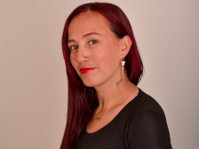 ChloeZenit has straight, deep red hair that falls past her shoulders. She wears a black, long-sleeved top and has red lipstick. Her right ear has a gold, winged earring, and a tattoo is visible on her right collarbone. She has a small nose ring in her right nostril. Her skin is fair, and her expression is neutral. The image is a simple portrait with a plain, light gray background. Her hair is glossy and her eyes are brown.