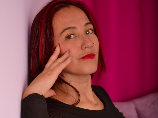 ChloeZenit with red and black hair, nose piercing, red lipstick, and a black top, touching her cheek with a hand. She has a tattoo on her neck.