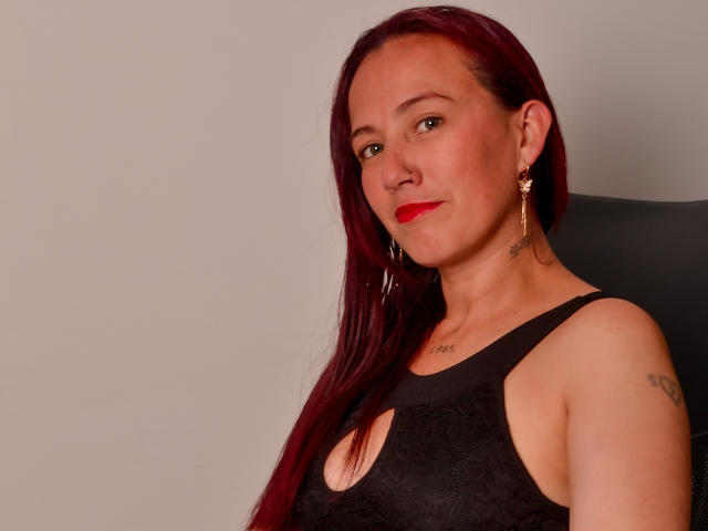 ChloeZenit has red hair, red lipstick, black dress with cutout, earrings, tattoo on right shoulder, and a confident, slightly smirking face. She's sitting against a dark chair.
