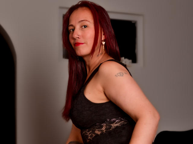 ChloeZenit has red hair, a tattoo on her right arm, wearing a black lace tank top, and red lipstick, standing with a confident, slightly smug expression.
