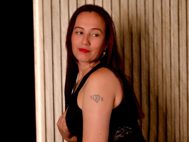 ChloeZenit has red lipstick, long brown hair, and a black lace top. She's holding her tits with a heart tattoo on her right arm, smiling slightly.
