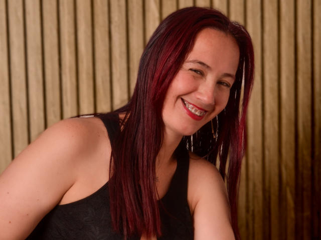 ChloeZenit, a woman with medium-length, straight, dark red hair, smiles broadly with visible white teeth. She wears a black sleeveless top and small, silver hoop earrings. Her skin is light, and she has a slight, natural-looking makeup with red lipstick. The image focuses on her upper body and face. The background consists of vertical, golden, wooden slats. The photo has a warm, indoor lighting. The composition is centered, and ChloeZenit's relaxed, happy expression is prominent.