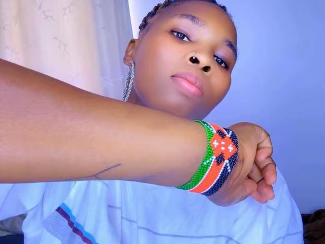 NattySlim, a young girl with dark brown skin and braided hair, has a nose ring and wears a chain earring. She has a serious expression and looks slightly to the side. She wears a white shirt with blue and red stripes. Her right arm is extended, showing a colorful bracelet with green, orange, black, and white stripes on her wrist. The bracelet has small white dots. A small tattoo is visible on her inner forearm. The light is blue, highlighting her skin tone.