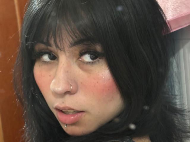 GaiaJoness has black hair, a nose piercing, and a lower lip piercing with a ball on her pink lips. Her eyes are looking to the side with a slight open-mouth expression.