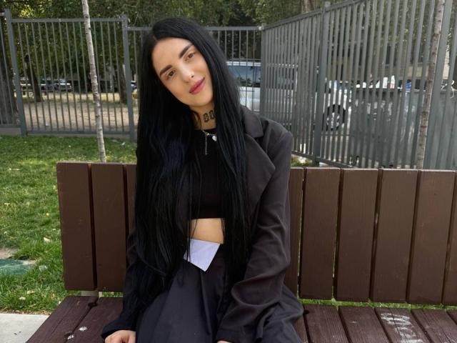 TiffanyHotxx with long black hair, black crop top, black coat, and black skirt, sitting on a brown wooden bench, looking at the camera with a slight smile.