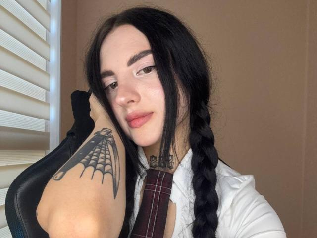 TiffanyHotxx with long black hair in braids, wearing a white shirt and maroon tie, has a spider tattoo on her arm, sitting in a chair, smiling softly.