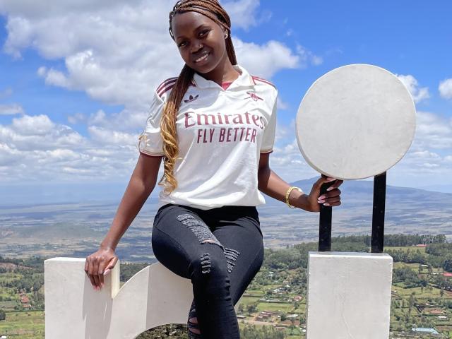 Sashie with dark skin, long braided hair, wearing a white Emirates Fly Better shirt and ripped black jeans, holding a round sign, smiling, sitting on a white structure.