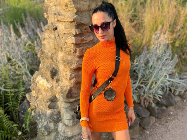 RubyValentine-hot with black hair in a ponytail, wearing an orange dress, brown shoulder bag, and sunglasses, standing against a palm tree, showing tattoos on her right arm.