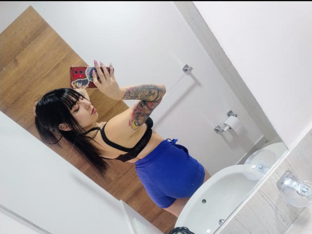 KuroBabee with black bra, blue shorts, and tattoos, taking a mirror selfie, showing her ass, holding a red phone with a sticker. Her arm tattoo is visible.