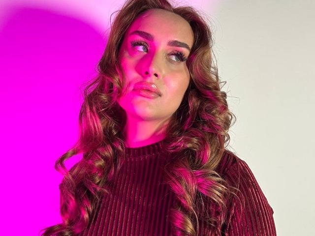 DawnGwin with curly brown hair, pink highlight, wearing a ribbed maroon sweater, looking up with a slight pout, pink light on the left side.