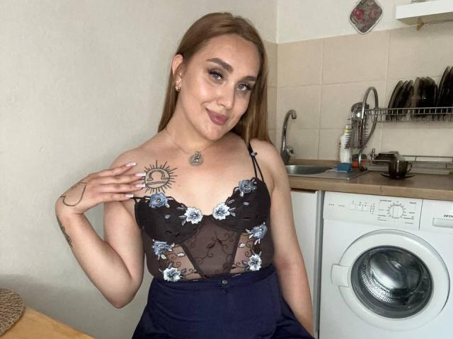 DawnGwin with long brown hair, wearing a black lace top with blue floral embroidery, sun tattoo on right shoulder, smiling, standing in a kitchen with a washing machine.