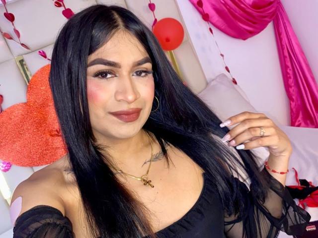 SilvanaCastro with long black hair, wearing a black off-shoulder top, showing her tits, pink blush, hoop earrings, gold necklace, ring, and pink lipstick, smiling at the camera.
