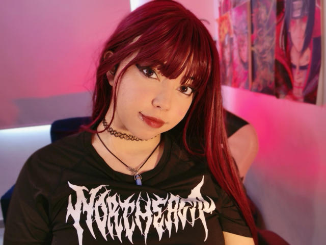 Arieyaara, con cabello rojo, un collar negro y una camisa de estilo gótico, mostrando sus pechos, tiene una cara linda y un pequeño piercing en la nariz, y está sentada en una habitación iluminada con luz rosa..