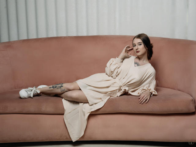 AlexLovely with tits visible, lying on a pink sofa in a beige dress, white shoes, tattoos on left leg, brown hair, red lipstick, hand touching face, relaxed pose.