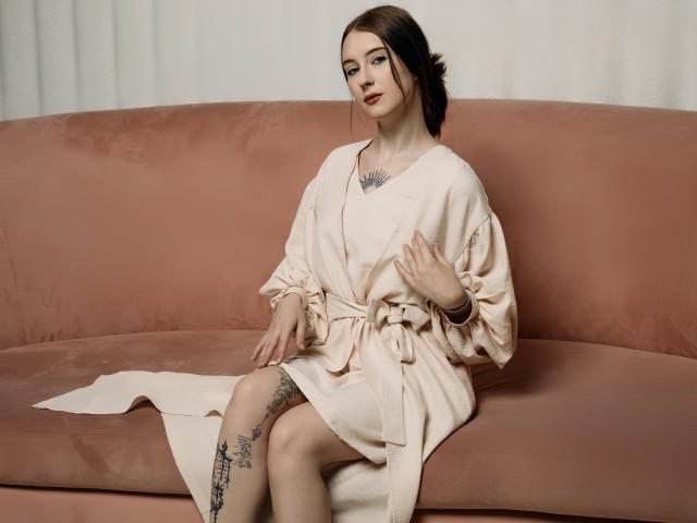 AlexLovely sits on a pink suede sofa, wearing a loose, beige robe tied at the waist. She has a tattoo on her left thigh, featuring intricate black designs. Her dark brown hair is pulled back, and she has a subtle, confident expression. A small necklace with a black geometric pattern rests on her chest. Her right hand is slightly raised, and her left hand rests on her knee. The robe's fabric is slightly wrinkled, adding texture to the image. The overall composition is balanced and elegant.