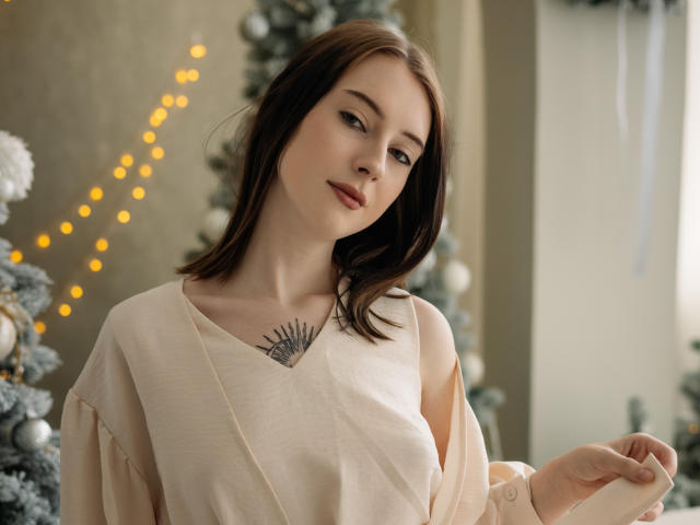 AlexLovely with brown hair, pale skin, small tits, wearing a beige top, showing a hand tattoo, and a gentle smile, in a softly lit, Christmas-decorated room.