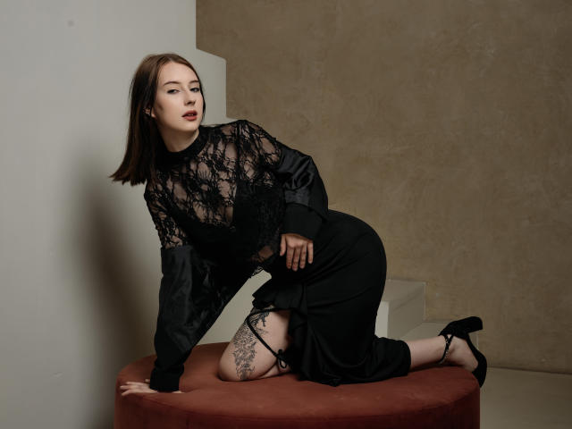 AlexLovely, kneeling on a red cushion, wearing a black lace top and skirt, showing thigh tattoos, with a serious expression, and black high heels.