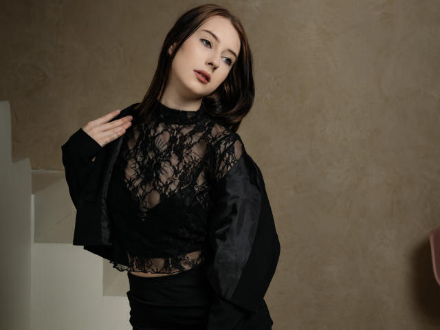 AlexLovely with dark brown hair, wearing a black lace top showing her tits, and a black jacket, standing against a beige wall, looking at the camera with red lipstick.