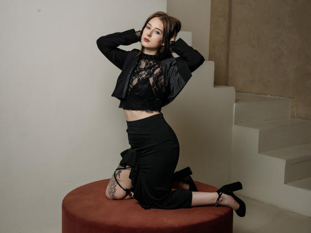 AlexLovely kneeling on a red circular cushion, wearing a black lace crop top, black jacket, black high-waisted skirt, black lace stockings, and black high heels, with hands in her brown hair.
