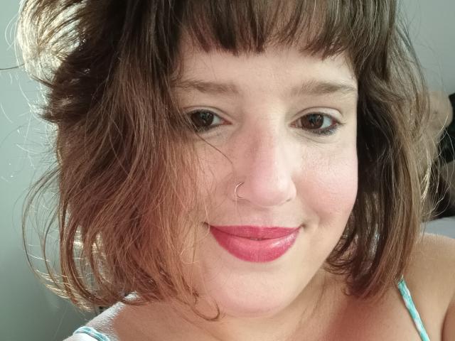Mmary hat braunes, welliges Haar, einen Nasenring und pinken Lippenstift. Ihre Brüste sind nicht sichtbar. Sie lächelt mit rotem Lippenstift. Ihre Augen sind braun. Ihre Haut ist hell..