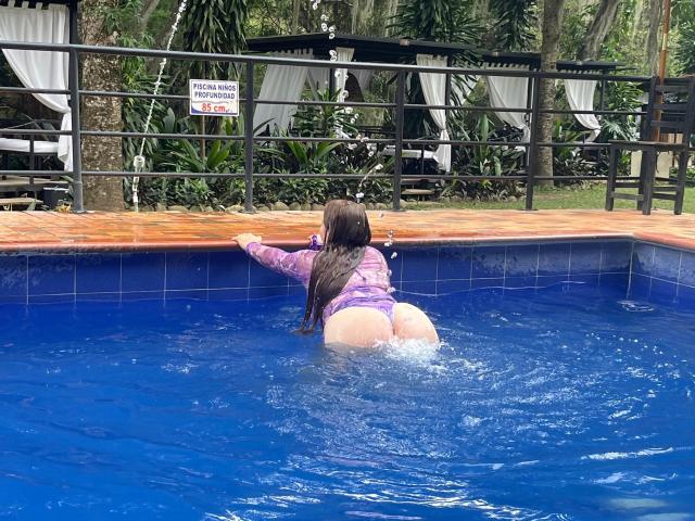 ValentinaDiazHot with wet butt in blue pool, wearing purple top, kneeling with back to camera, hands on edge, brown hair, water splashing, tropical setting.