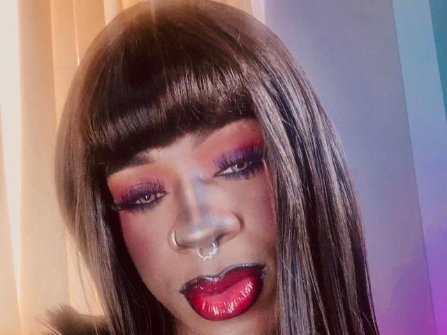 EbonyHansNight has shiny black hair, bright red lipstick, purple eyeshadow, thick eyelashes, and a nose ring, looking down with a slightly open mouth.