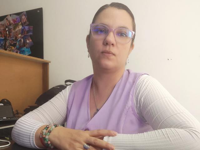Amelia Wilson, con gli occhiali, indossa un gilet color lavanda chiaro, una maglietta bianca a coste, ha i capelli castani raccolti in una coda di cavallo, braccialetti verdi e un anello, appare leggermente infastidita e siede a una scrivania..